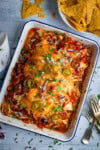 A baking dish filled with cheesy chicken salsa bake, topped with jalapeños, red onions, and chopped herbs, with a bowl of tortilla chips and cutlery nearby on a light surface.