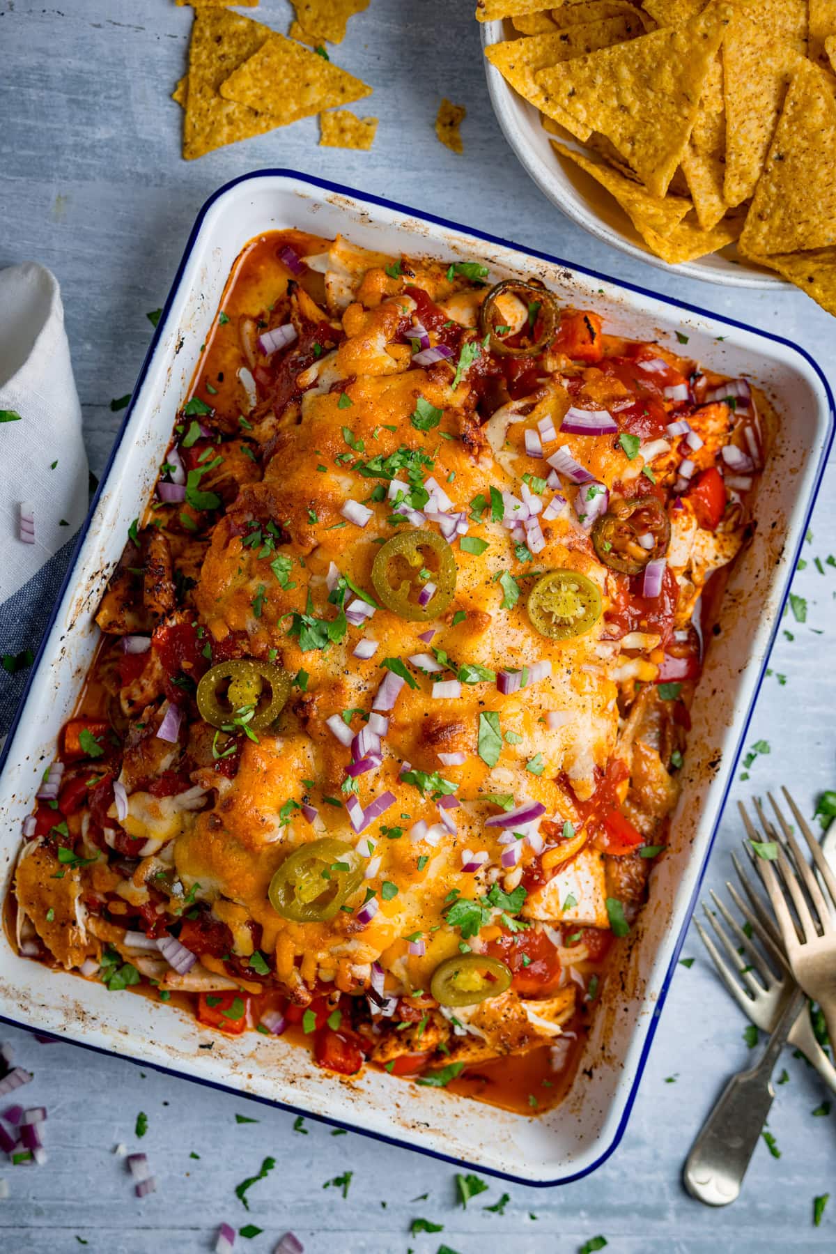 A baking dish filled with cheesy chicken salsa bake, topped with jalapeños, red onions, and chopped herbs, with a bowl of tortilla chips and cutlery nearby on a light surface.