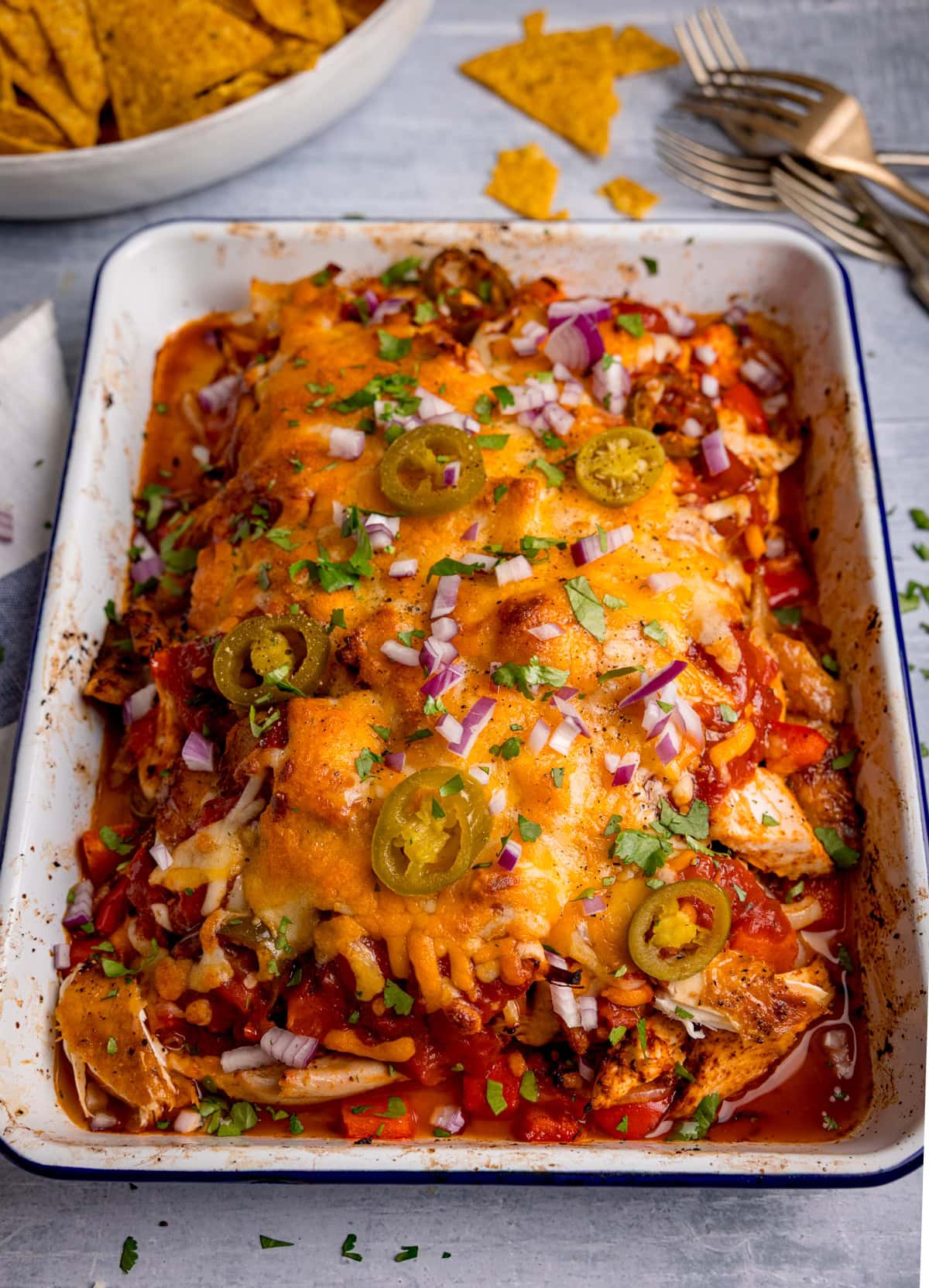 A baking dish filled with a salsa chicken bake, topped with melted cheese, jalapeños, diced red onions, chopped coriander, red peppers, and tomato sauce.
