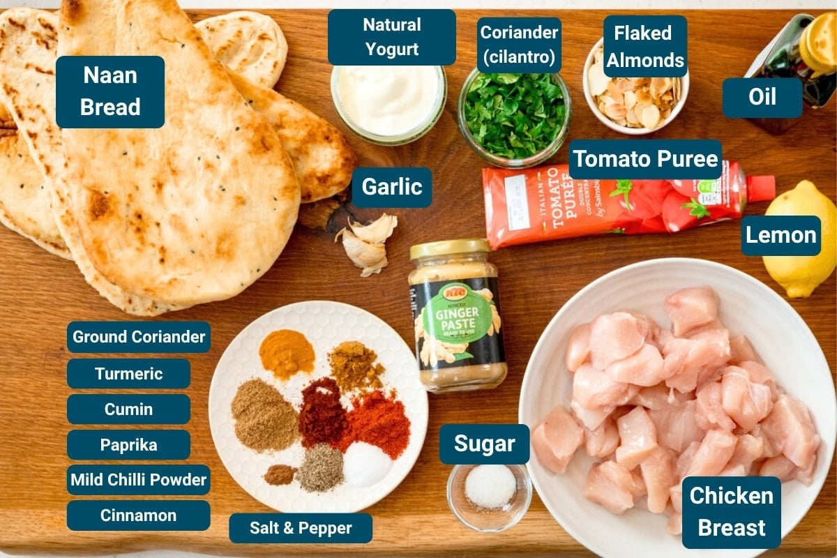 A wooden board with labelled ingredients for naan bread chicken pizza including naan bread, raw chicken, spices, ginger purée, tomato purée, yoghurt, parsley, flaked almonds, lemon, garlic, and seasonings.