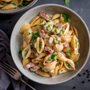 A bowl of creamy pasta with peas and bacon, featuring pasta shells, garden peas, parsley, and smoky bacon, served on a grey surface with a fork and serviette nearby.