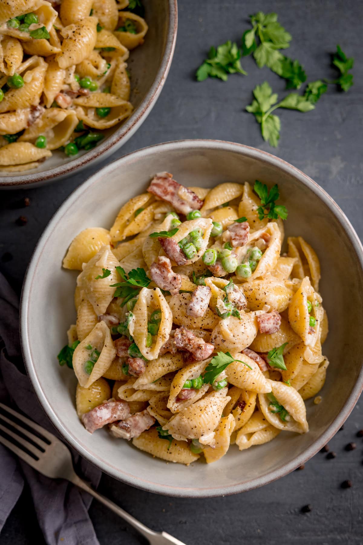 A bowl of pasta with bacon and peas, tossed in a creamy sauce with parsley, and finished with black pepper; another bowl and scattered parsley, with bacon and peas in the background.