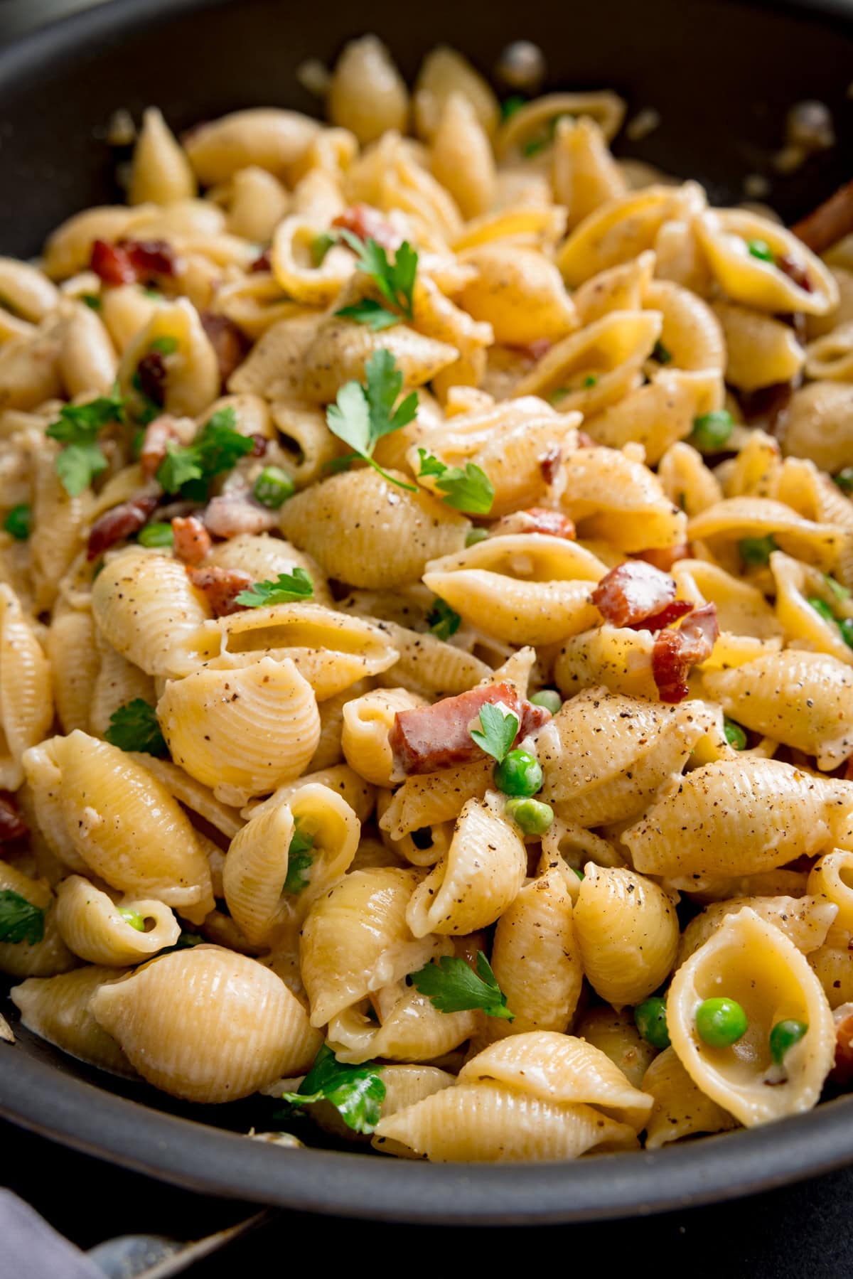 A frying pan of creamy pasta with peas and bacon, tossed with parsley in a creamy sauce and topped with freshly ground black pepper.