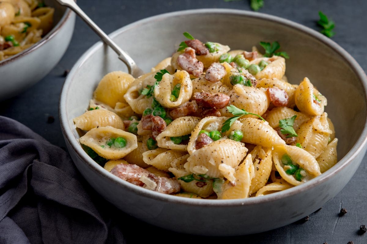 A bowl of creamy pasta with peas and bacon, mixed in a creamy sauce and garnished with parsley, with a fork resting inside the bowl.
The bowl is on a grey background, next to a grey napkin, with a further bowl of pasta just visible in the background.