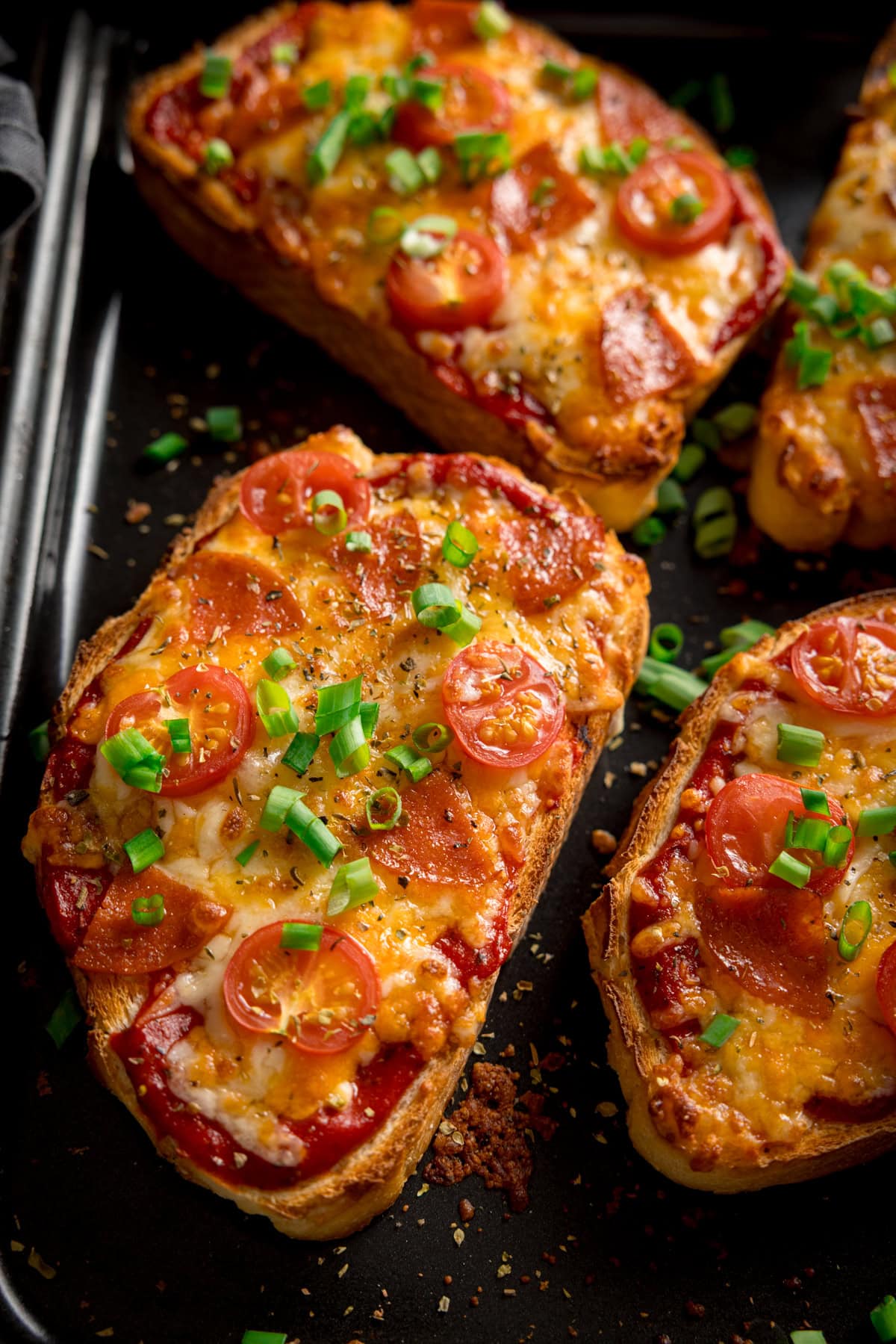 Rebanadas de pan cubiertas con queso derretido, pepperoni, tomates cherry y cebolletas picadas en una bandeja de horno negro.