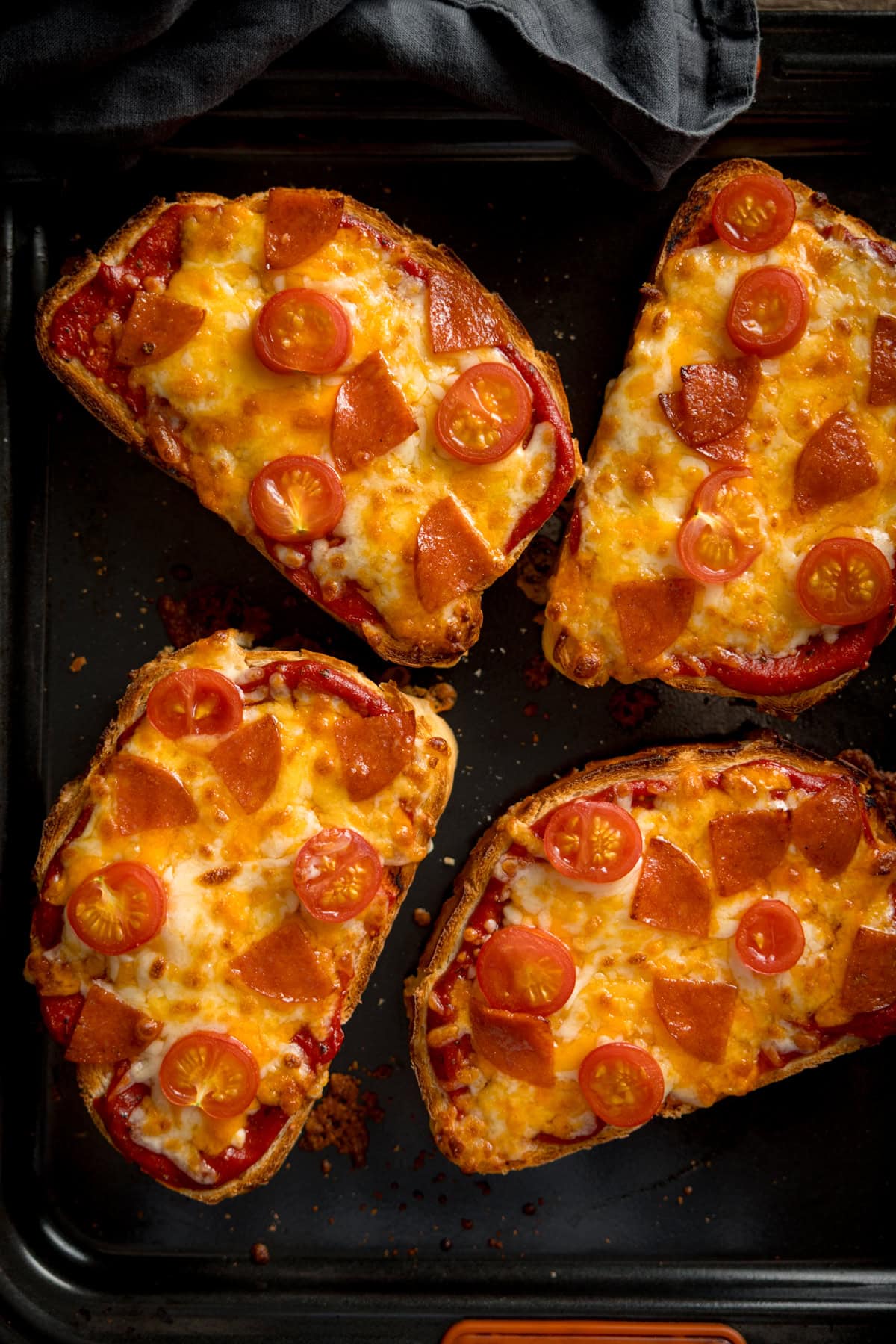Cuatro rebanadas de pan cubiertas con queso derretido, pepperoni en rodajas y tomates cherry a la mitad en una bandeja para hornear.