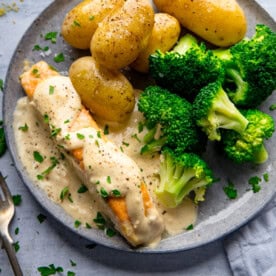 A plate with boiled potatoes, steamed broccoli, and pan fried salmon fillet topped with a creamy white sauce, garnished with chopped parsley.