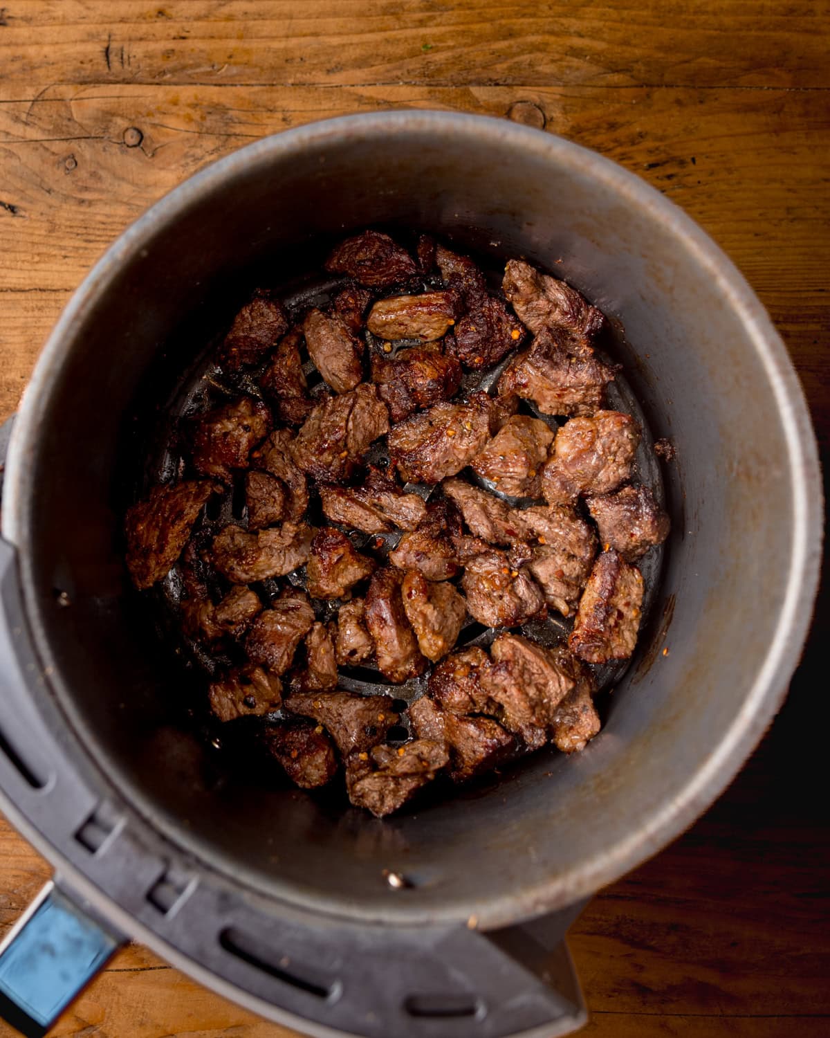 Cooked steak pieces are shown inside an air fryer basket, placed on a wooden surface.