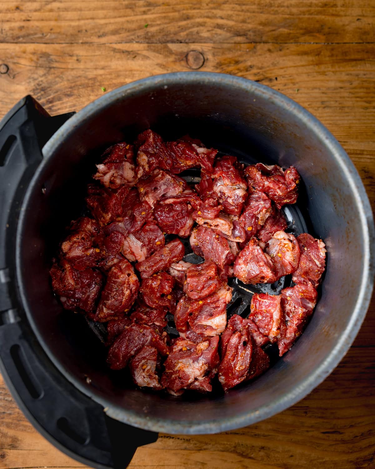 Chunks of raw, seasoned steak, for making Air Fryer Steak Bites inside an air fryer basket on a wooden surface.