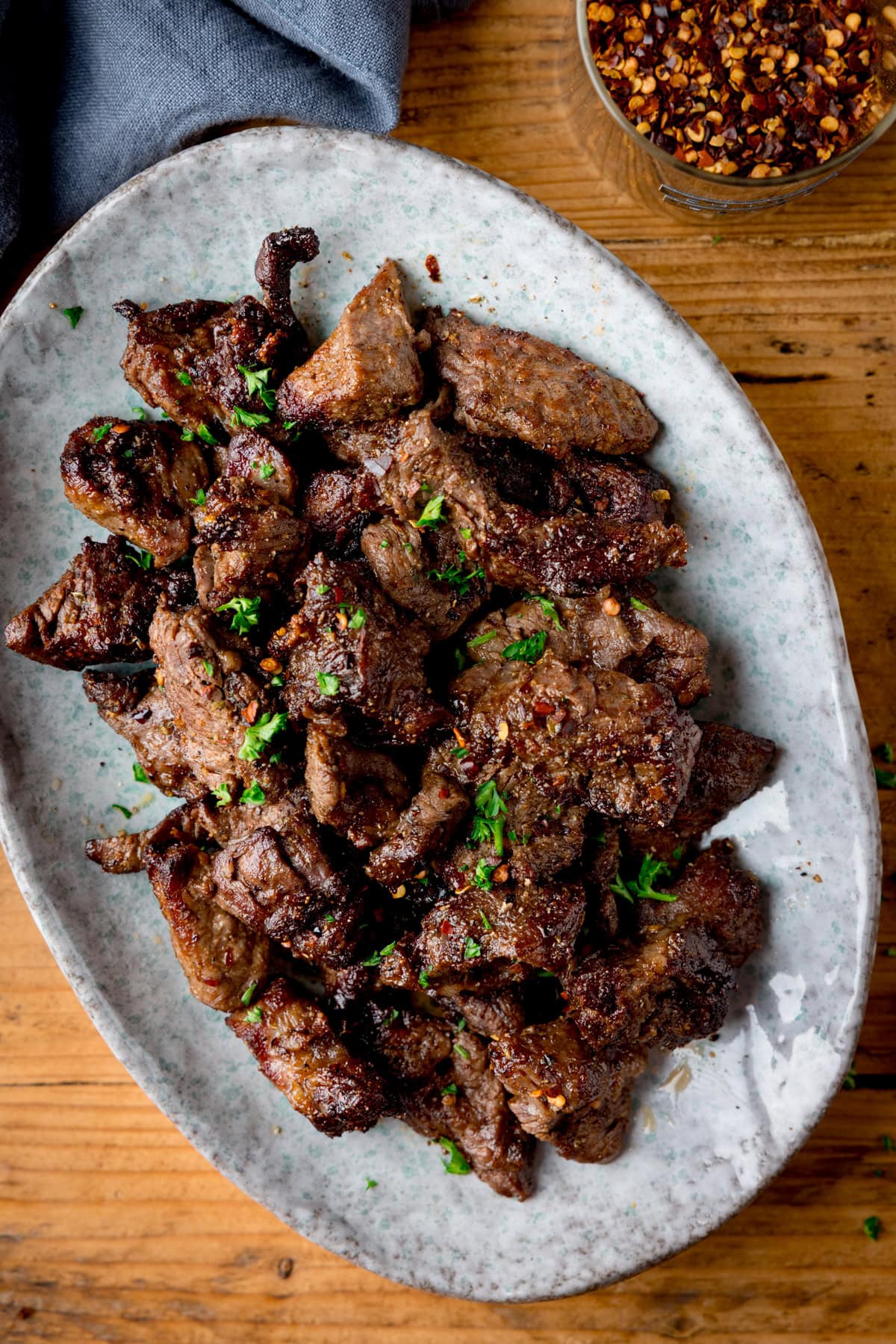 A light oval plate filled with seasoned steak bites garnished with chopped parsley, placed on a wooden table.
