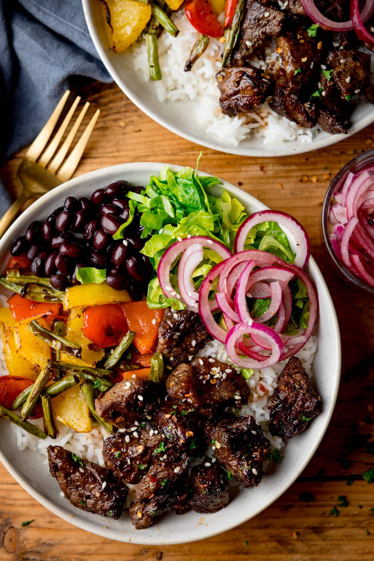 A bowl of rice topped with Air Fryer steak pieces, sliced red onions, black beans, lettuce, and roasted peppers, with a gold fork and another bowl partially visible.
The bowls are on a wooden table.