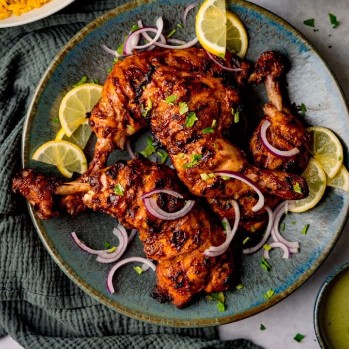 Tandoori chicken legs grilled to perfection, garnished with lemon slices, red onion, and chopped herbs on a blue plate.