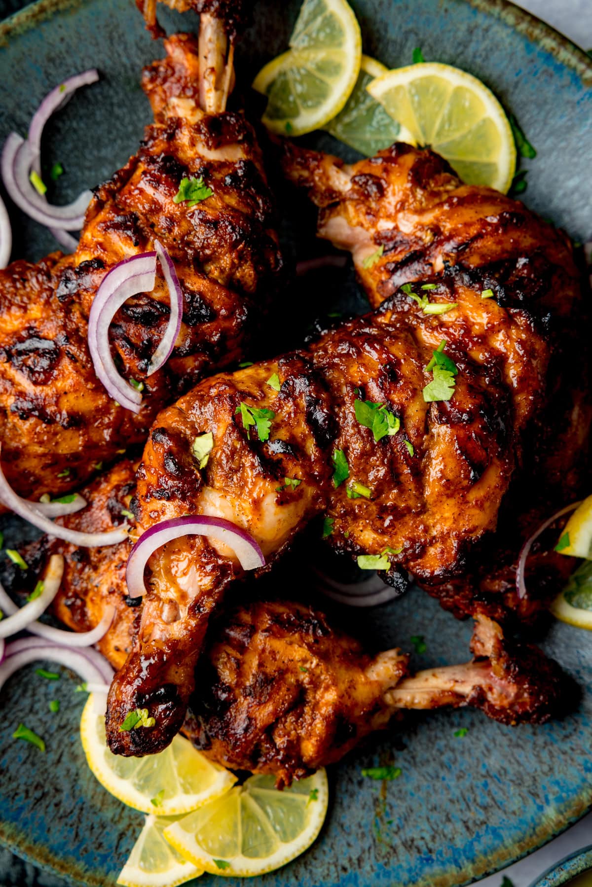 Grilled tandoori chicken legs on a blue plate. garnished with sliced red onions, fresh coriander, and lemon wedges.