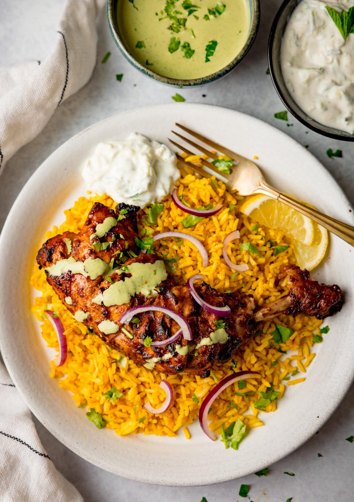 A plate of yellow rice topped with succulent tandoori chicken, red onion slices, chopped herbs, lemon wedges, and a dollop of raita, with extra sauces in bowls nearby.