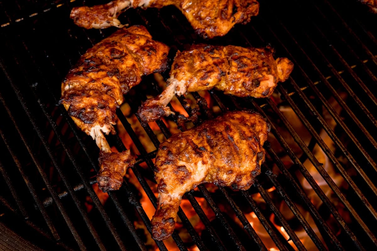 Four pieces of tandoori chicken with char marks are cooking on a barbecue over open flames.