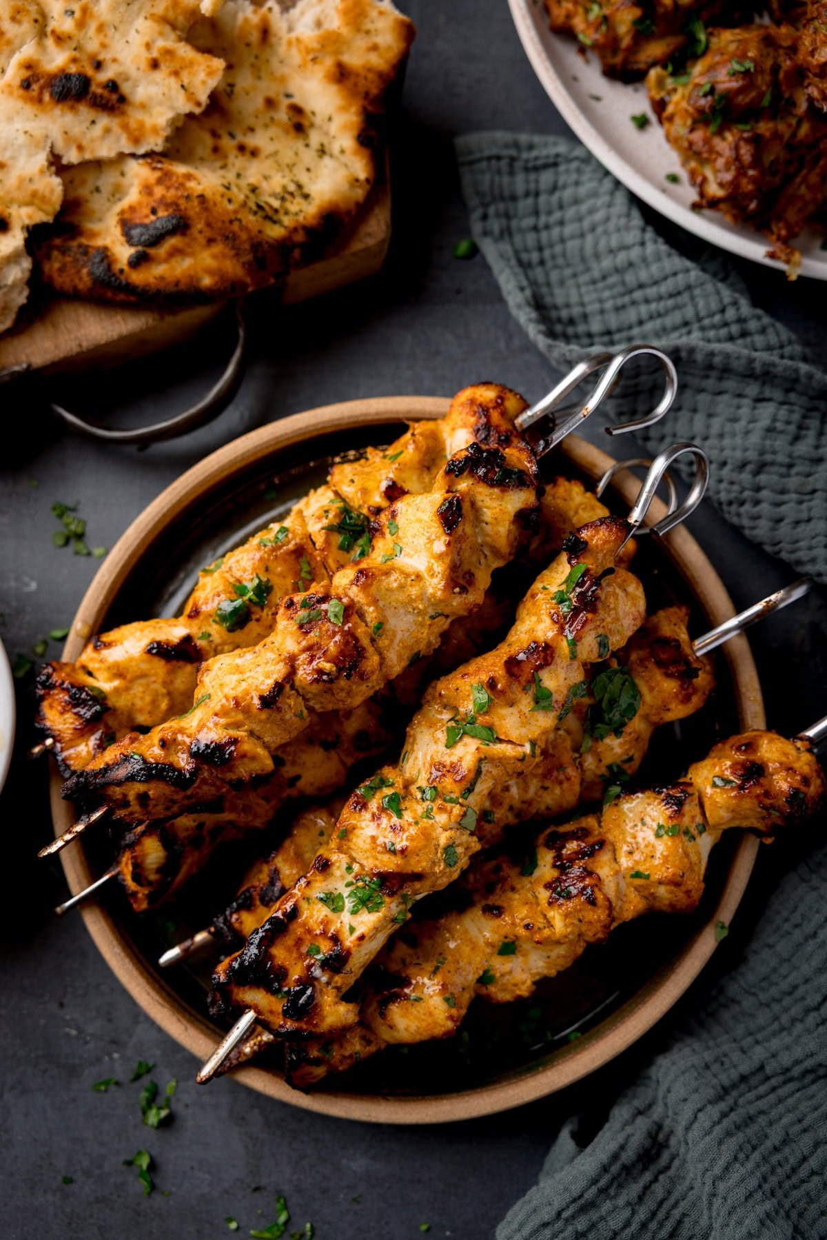 Skewers of Air Fryer Chicken Tikka garnished with fresh cilantro on a plate. Pieces of naan and a green napkin are nearby.