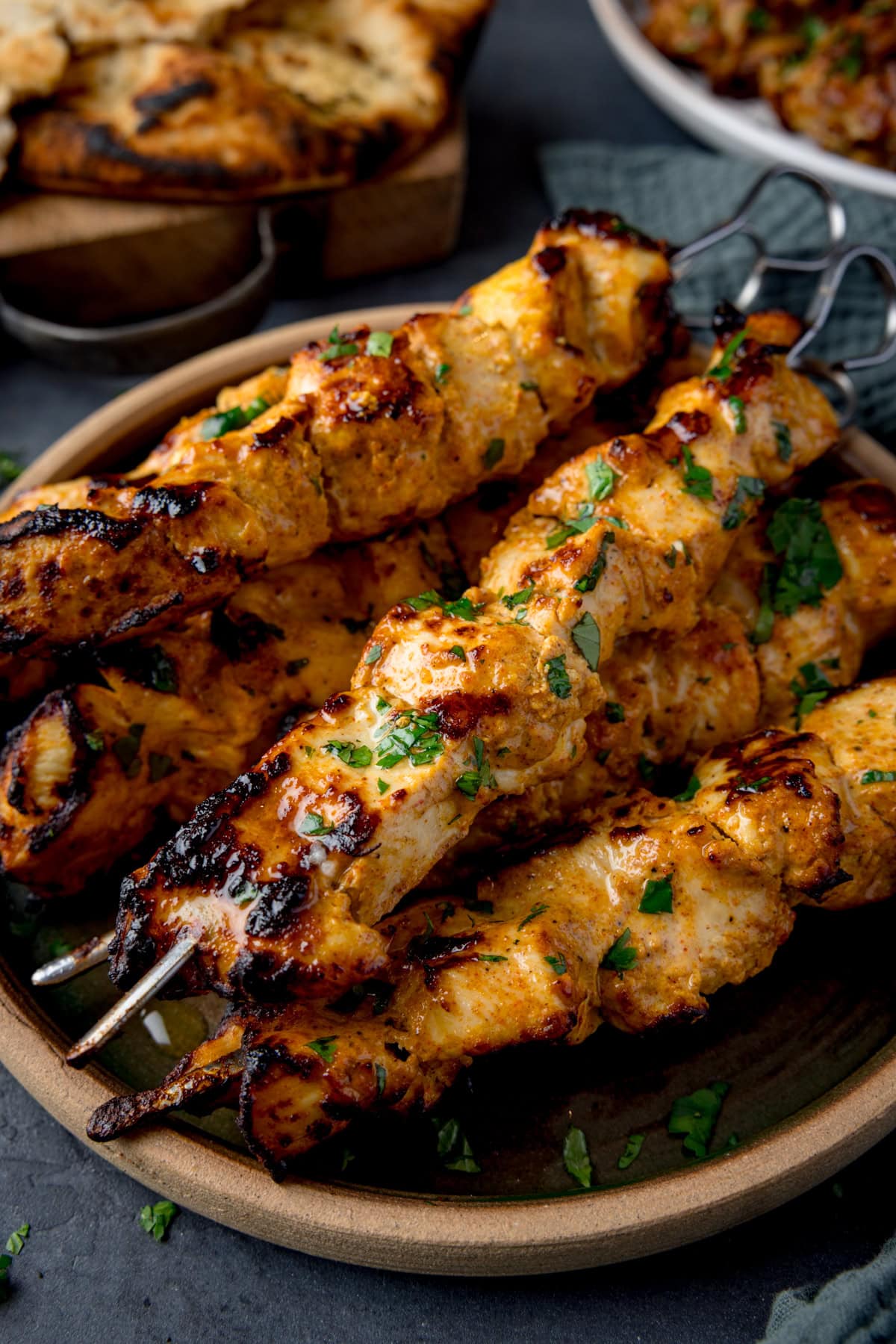 Close up of Air Fryer Chicken Tikka skewers garnished with chopped cilantro on a plate. Naan bread is visible on a board at the top of the frame.