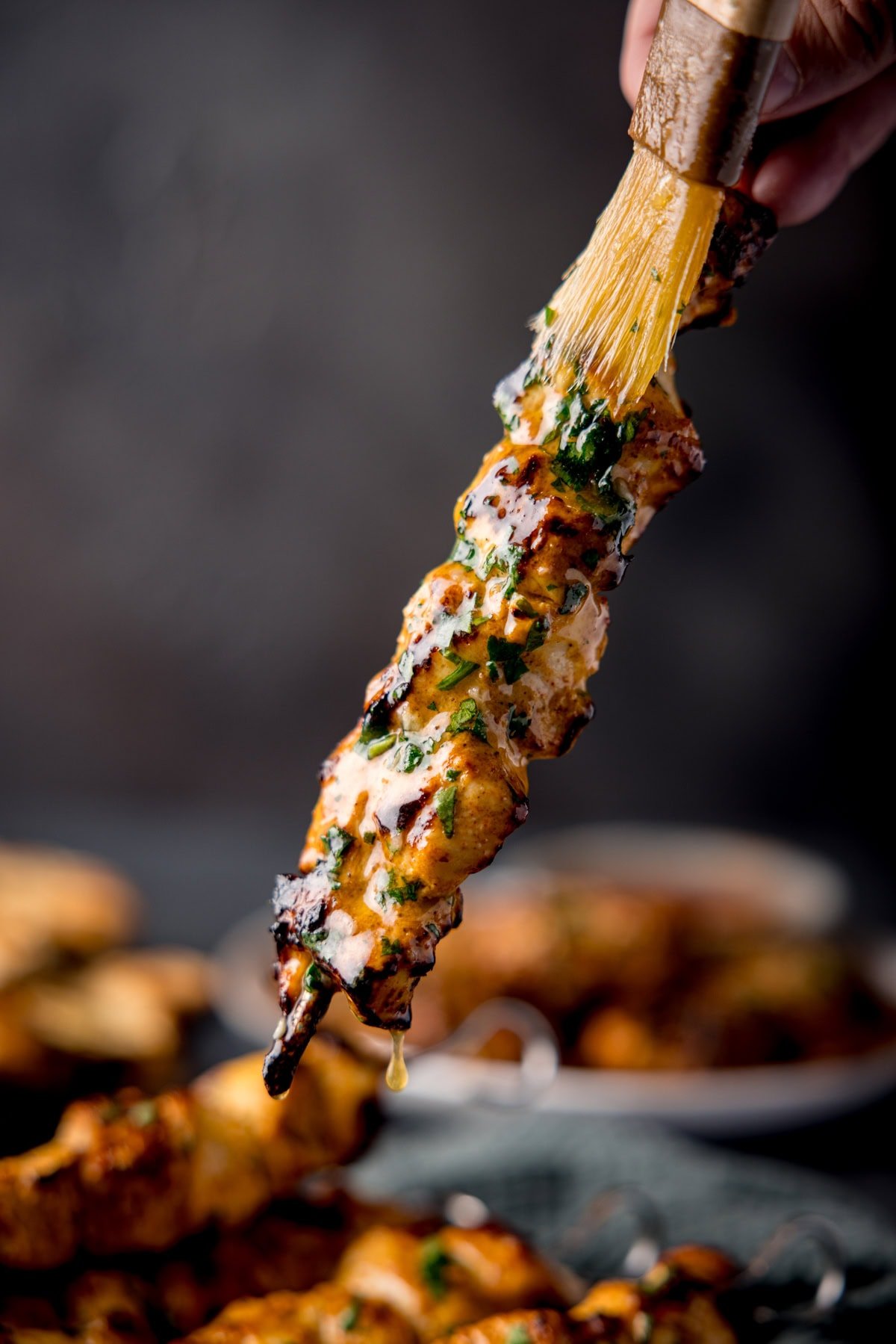 A skewer of Chicken Tikka being brushed with cilantro garlic butter. More skewers are blurred in the background.