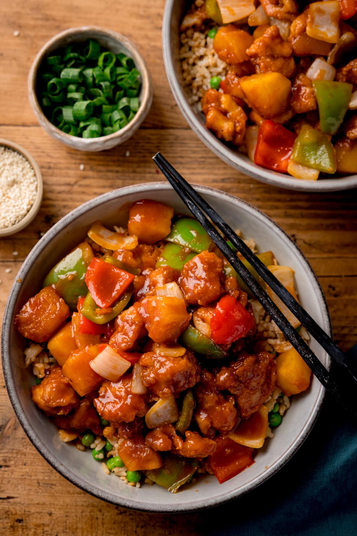 Tall overhead image of a bowl of sweet and sour chicken with chunks of onion, red & green peppers and pineapple in sauce. The bowl is on a wooden surface and a pair of black chopsticks rest on the bowl. A further bowl of sweet and sour chicken is partly in shot at the top of the frame. Small bowls of chopped green onion and sesame seeds are nearby.