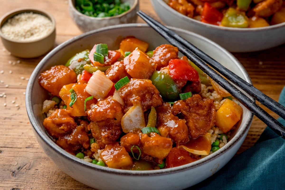 Wide side-on image of a bowl of sweet and sour chicken with chunks of onion, red & green peppers and pineapple in sauce. The bowl is on a wooden surface and a pair of black chopsticks rest on the bowl. A further bowl of sweet and sour chicken is partly in shot at the top of the frame. Small bowls of chopped green onion and sesame seeds are nearby.