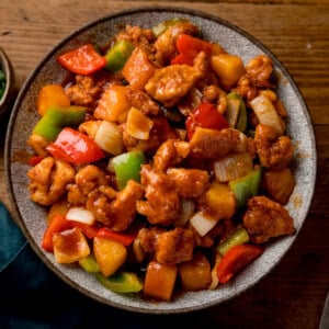 Square overhead image of a bowl of sweet and sour chicken with chunks of onion, red & green peppers and pineapple in sauce. The bowl is on a wooden surface.