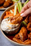 A buffalo chicken wing is being dipped into a small bowl of blue cheese sauce. These are celery and carrot sticks also on the plate in the background.