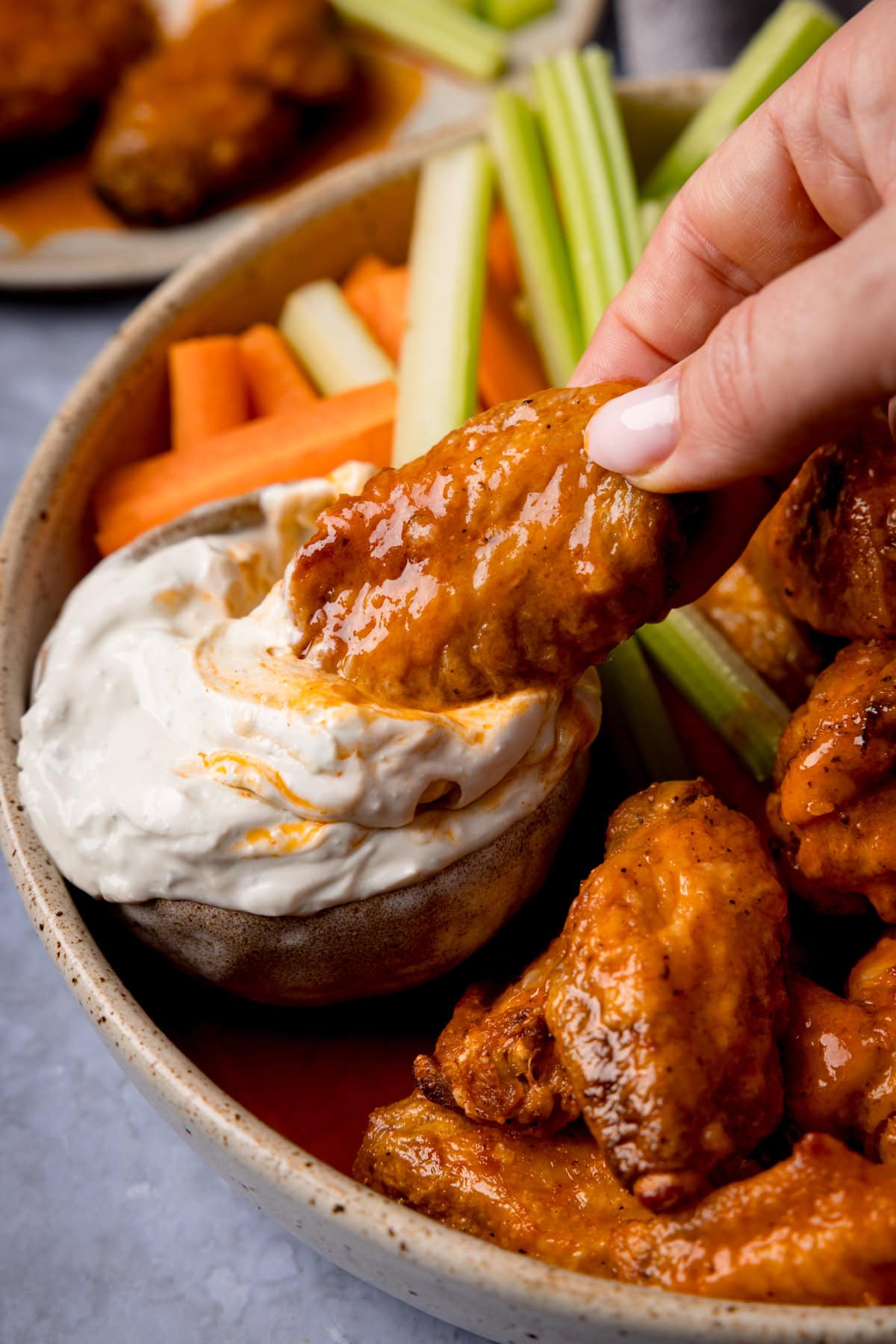 A buffalo chicken wing is being dipped into a small bowl of blue cheese sauce. These are celery and carrot sticks also on the plate in the background.