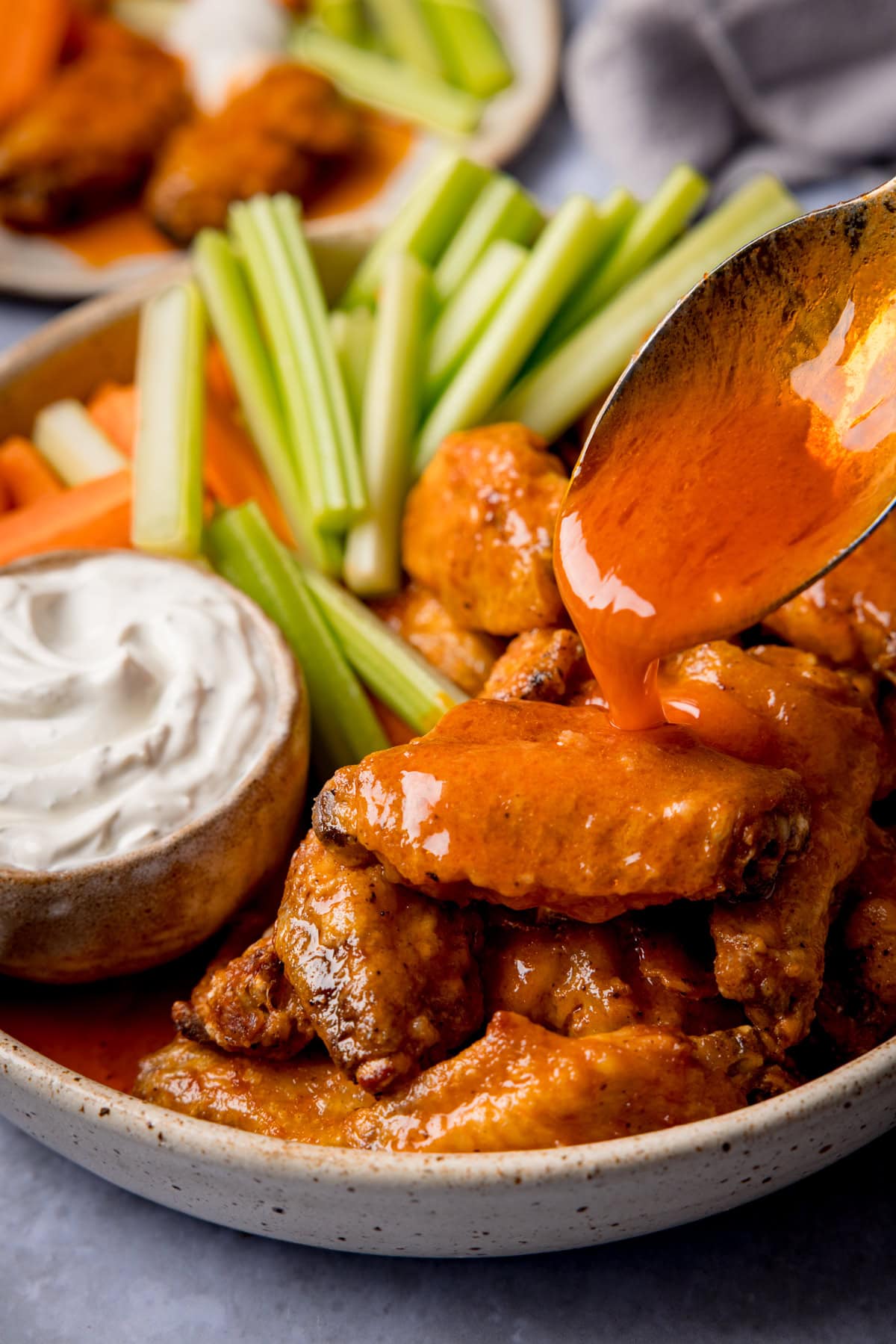 A plate of spicy air fryer wings, served with celery, carrot batons and blue cheese dip. Buffalo sauce is being poured over one of the wings.