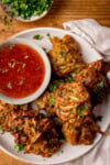 A plate of crispy Air Fryer onion bhajis garnished with chopped coriander on a white plate. They're served with a small bowl of sweet chilli sauce. The plate is on a wooden table.