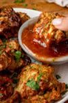 A crispy air fryer onion bhaji being dipped into a small bowl of sweet chilli sauce. There are more onion bhajis on the plate, sprinkled with chopped coriander.