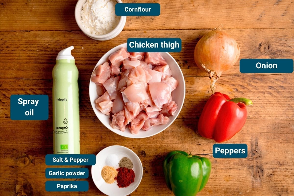 Chopped chicken thigh in a bowl, cornflour, spray oil, onion, red and green peppers, and spices on a wooden table. Each item is labelled.