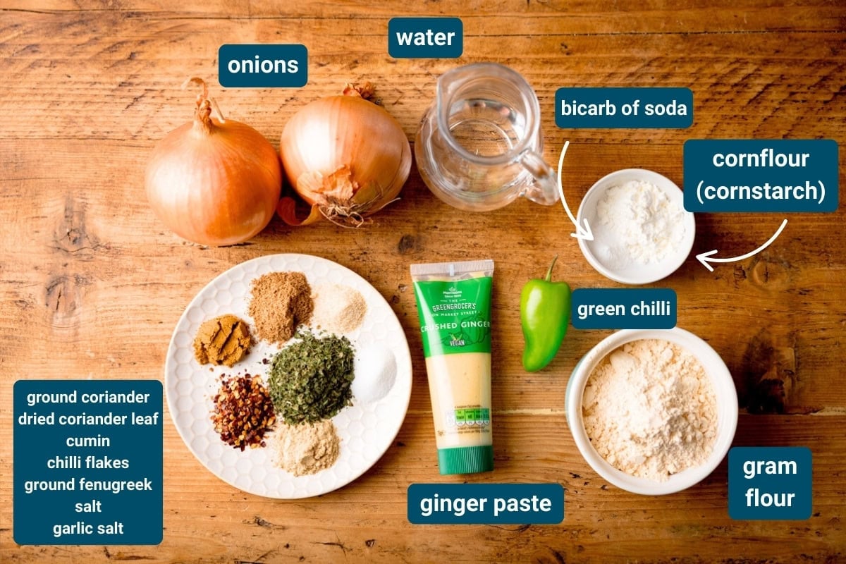 Ingredients for an air fryer Onion Bhaji recipe arranged on a wooden surface: onions, water, gram flour, cornflour, green chilli, ginger paste, and spices.