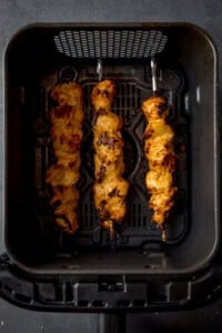 Three skewers of cooked Chicken Tikka sizzle in an air fryer basket, viewed from above.