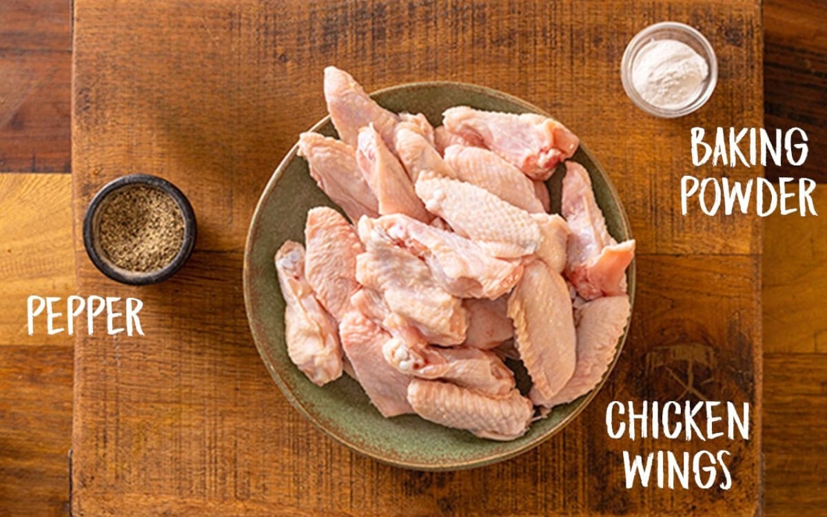 A bowl of raw chicken wings on a wooden table next to a small bowl of pepper and a small bowl of baking powder.