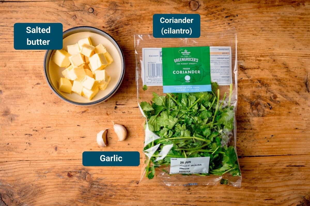 Ingredients Cilantro Garlic Butter on a wooden table. The ingredients are labelled, these are: a bowl of cubed salted butter, two garlic cloves, a pack of fresh coriander.
