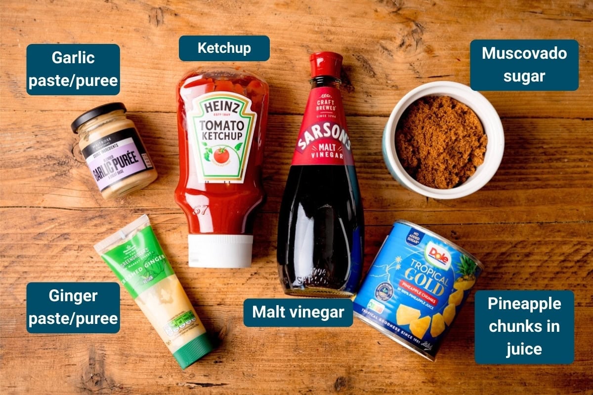 Six ingredients on a wooden surface, including garlic paste, ginger paste, tomato ketchup, malt vinegar, muscovado sugar, and tinned pineapple in juice. Each item is labelled.