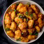 Close-up overhead square image of a white bowl filled with crispy Air Fryer hot honey chicken bites, garnished with chopped spring onions and sesame seeds.