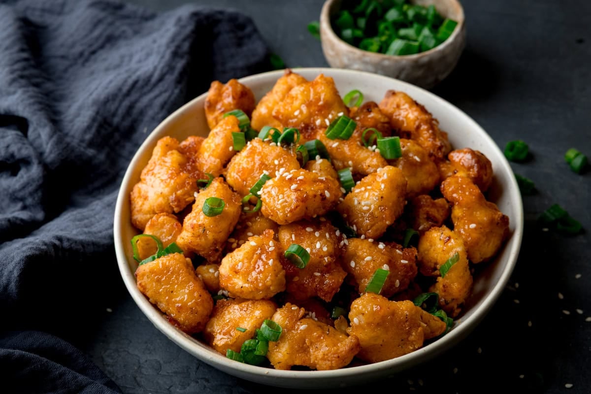 A bowl of crispy chicken pieces coated in hot honey and sprinkled with sesame seeds.