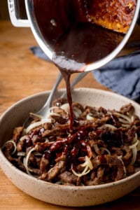 Chilli beef sauce is being poured over a bowl of crispy chilli beef strips and sliced onions on a wooden table.