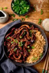 A black bowl filled with air fryer crispy chilli beef with onions, sesame seeds, and sliced spring onions, served with fried rice. The bowl is on a wooden surface next to a blue/grey napkin, a pair of chopsticks, a small jug of soy sauce, a small bowl of chopped spring onions and a small dish of sesame seeds.