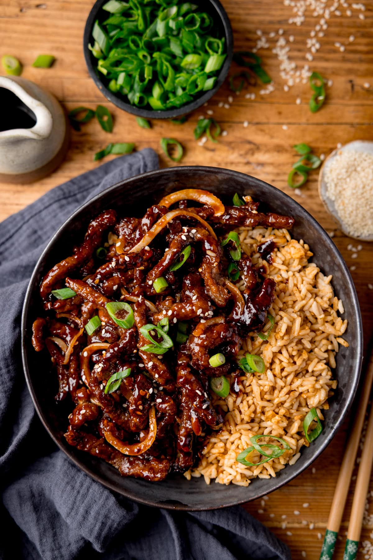 A black bowl filled with air fryer crispy chilli beef with onions, sesame seeds, and sliced spring onions, served with fried rice. The bowl is on a wooden surface next to a blue/grey napkin, a pair of chopsticks, a small jug of soy sauce, a small bowl of chopped spring onions and a small dish of sesame seeds.