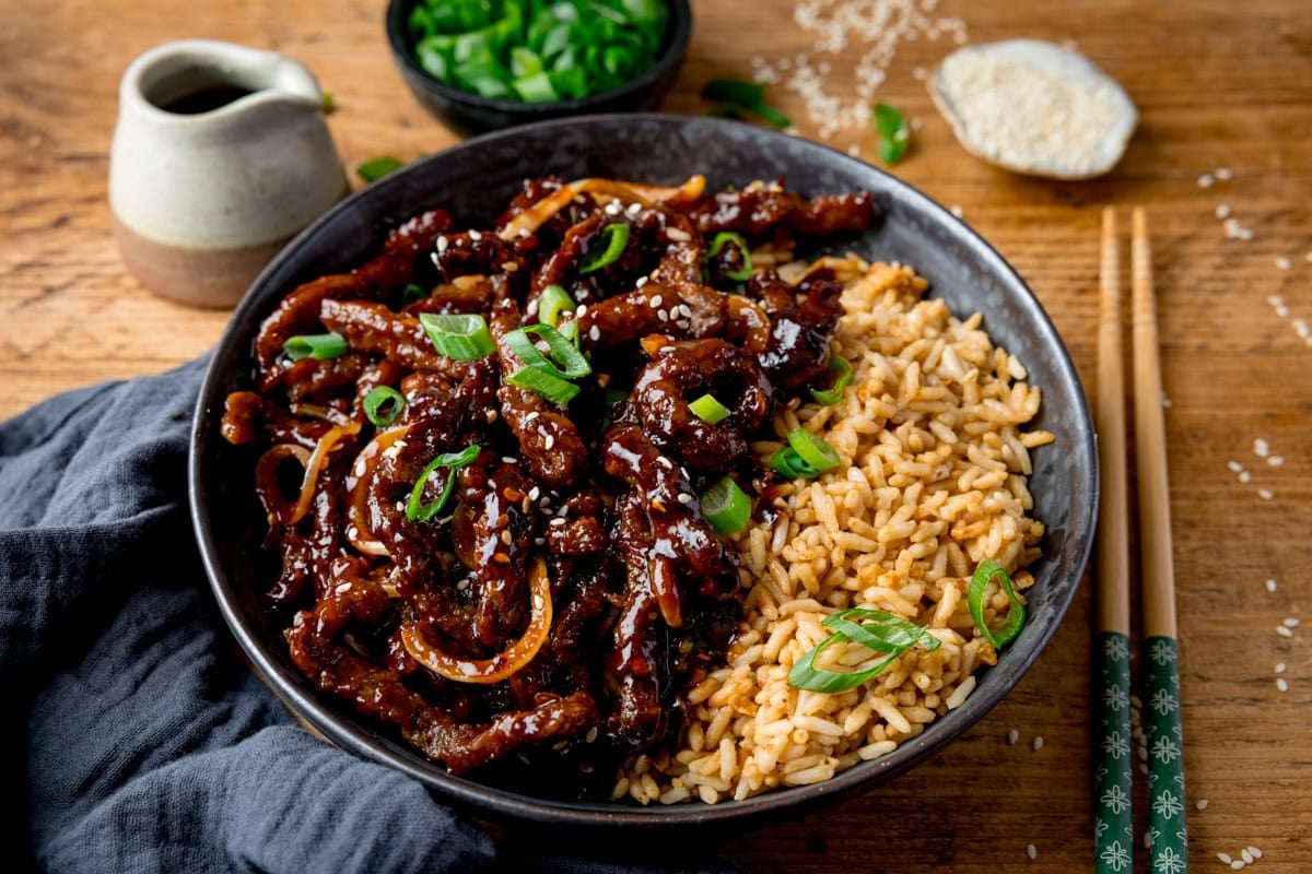 A wide image of a black bowl filled with air fryer crispy chilli beef with onions, sesame seeds, and sliced spring onions, served with fried rice. The bowl is on a wooden surface next to a blue/grey napkin, a pair of chopsticks, a small jug of soy sauce, a small bowl of chopped spring onions and a small dish of sesame seeds.
