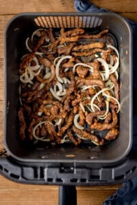 Strips of crispy beef and cooked onion slices in an air fryer basket, viewed from above.