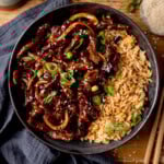A black bowl filled with air fryer crispy chilli beef with onions, sesame seeds, and sliced spring onions, served with fried rice. The bowl is on a wooden surface next to a blue/grey napkin, a pair of chopsticks and a small dish of sesame seeds.