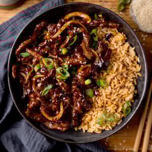 A black bowl filled with air fryer crispy chilli beef with onions, sesame seeds, and sliced spring onions, served with fried rice. The bowl is on a wooden surface next to a blue/grey napkin, a pair of chopsticks and a small dish of sesame seeds.