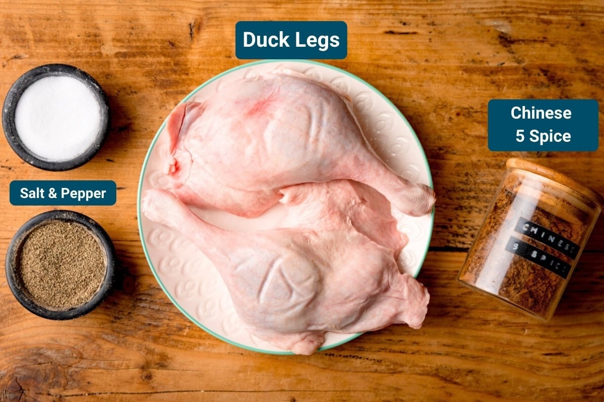 Ingredients for air fryer crispy duck on a wooden surface. The ingredients are labelled. They are: Two raw duck legs on a plate, pots of salt and pepper, a jar of Chinese 5 spice.