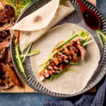 A metal plate with crispy duck slices, cucumber strips, and hoisin sauce on an open Chinese pancake. A rolled pancake and a wooden spoon with hoisin sauce on is also on the plate. The plate is on a wooden board and there is more shredded duck on the board.