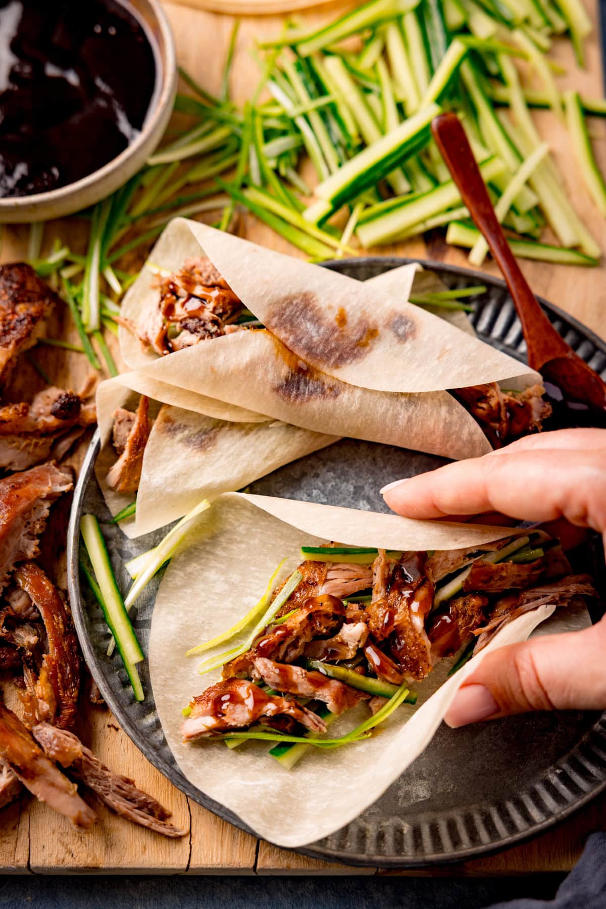 A hand is holding an open Chinese duck pancake with hoisin sauce and julienned spring onion and cucumber. The pancake is on a silver plate and there is also a rolled duck pancake and a wooden spoon with hoisin sauce resting on the plate. The plate is on a wooden board with shredded duck, more julienned vegetables and a dish of hoisin sauce nearby.