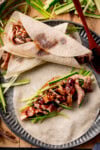 A plate with an open Chinese duck pancake with hoisin sauce and julienned spring onion and cucumber. There is also a rolled duck pancake and a wooden spoon with hoisin sauce resting on the plate. The plate is on a wooden board with more julienned vegetables nearby.