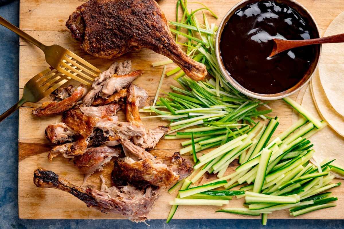 A wooden board with crispy duck, shredded next to two gold forks, sliced cucumber and spring onion, a bowl of hoisin sauce with a wooden spoon sticking out, and a pile of Chinese pancakes.